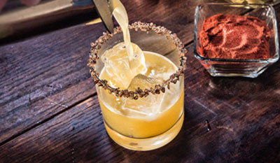 Yellow cocktail with brown rim being poured with a dish of a red powder next to it.