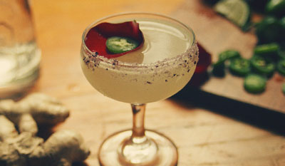 Close up of a margarita with a pink flower and jalapeño floating in it. Jalapeños and ginger are displayed in the background.