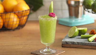 Frozen green margarita with a pink flower and celery garnish on a table with a basket of oranges, limes and celery in the background.