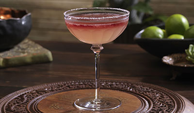 Pink cocktail with a salt rim and red floater on a brown table with a bowl of limes in the background.